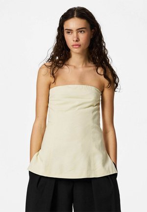 Kvinne med langt, krøllete hår iført en ermeløs beige tube-topp og løse svarte bukser, står med hendene i lommene mot hvit bakgrunn.