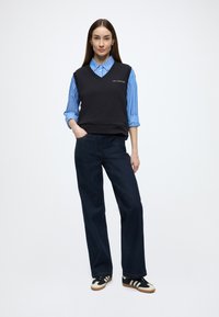 Camisola preta sem mangas sobre uma camisa de botões às riscas azuis e brancas, combinada com jeans escuros de perna larga e ténis pretos.