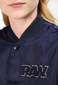 Námořnická modrá bomber bunda s žebrovaným límcem, zapínáním na knoflíky a vyšívaným logem "RAW." na hrudi. Hladký, lesklý textilní povrch.