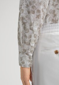 Helles graues Blümchenhemd mit langen Ärmeln und geknöpften Manschetten, kombiniert mit weißen Hosen, die eine Gesäßtasche und einen braunen Knopf haben.