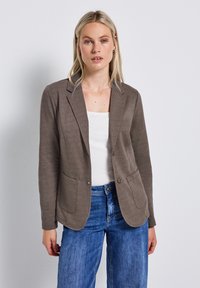 Blazer en tissu marron et doux avec deux poches avant et fermeture à un bouton, porté sur un haut blanc. Associe avec un jean en denim bleu.