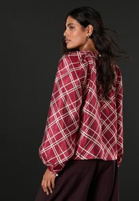 Kvinde med langt mørkt hår iført en skinnende rød bluse med hvidt rutenetmønster og voluminøse ærmer, poserende mod en mørk baggrund.