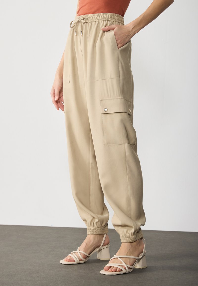 Beige cargo pants with a drawstring waist, elastic cuffs, side and cargo pockets, paired with light-colored strappy block-heeled sandals.