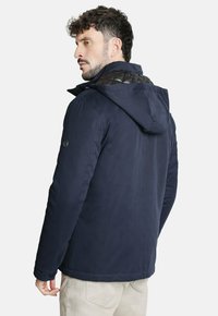 Chaqueta azul marino con cuello acolchado, capucha y detalles de botones. Presenta una textura suave y un corte ajustado, adecuado para el clima frío.