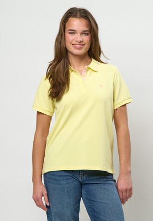 Junge Frau mit langen braunen Haaren, die ein hellgelbes kurzärmeliges Poloshirt und blaue Jeans trägt, lächelnd vor einem einfarbigen Hintergrund.