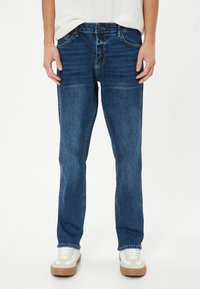 Jeans de mezclilla azul oscuro con un corte recto, detalles de costura, cinco bolsillos y cierre de botones, combinados con zapatillas blancas.