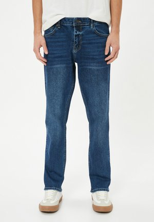 Jeans de mezclilla azul oscuro con un corte recto, detalles de costura, cinco bolsillos y cierre de botones, combinados con zapatillas blancas.