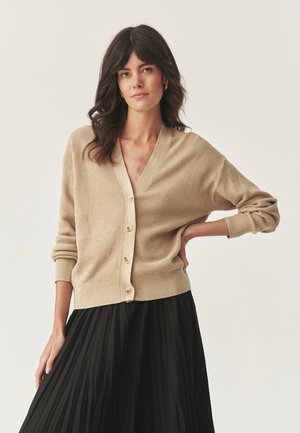 Donna con capelli scuri e mossi, che indossa orecchini d'oro, un cardigan beige con bottoni e una gonna nera plissettata, in piedi con una mano sul fianco.