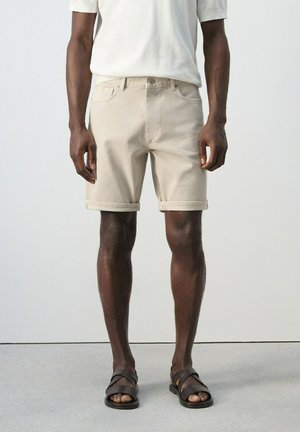 Hombre con pantalones cortos beige remangados, camisa blanca de manga corta y sandalias negras abiertas en los dedos, de pie frente a un fondo blanco sencillo.