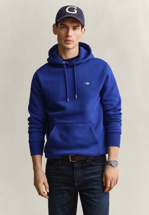 Junger Mann trägt einen blauen Hoodie, dunkle Jeans, Armbanduhr und eine marineblaue Kappe mit dem Buchstaben G, steht vor einem schlichten hellen Hintergrund.