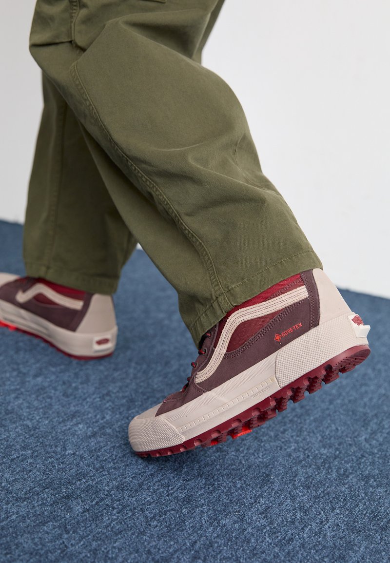 Weinrote und beige Sneaker mit strukturierten Gummisohlen und Gore-Tex-Akzenten; getragen mit olivgrünen Hosen auf einem blauen Teppich.