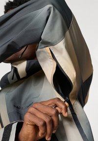 Person wearing color-blocked jacket with beige, gray, and black sections, pulling up a zipper on the sleeve near the wrist.