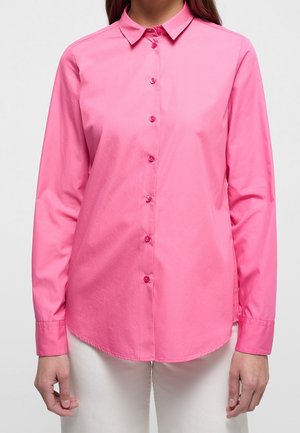 Femme portant une chemise rose vif à manches longues boutonnée avec col, associée à un pantalon blanc, debout devant un fond clair uni.