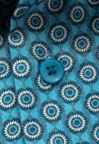 Blauer Stoff mit kreisförmigen Mustern in Beige und Dunkelblau, mit einer Nahaufnahme eines runden blauen Knopfes, der mit blauem Faden angenäht ist.