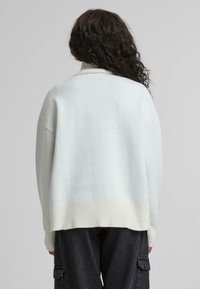 Hellgrauer, übergroßer Pullover mit geripptem Saum und Bündchen, der einen hohen Kragen hat. Die glatte Textur und der lässige Schnitt sind von hinten sichtbar.