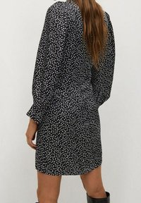 Abito nero con motivo floreale bianco, maniche lunghe e vestibilità ampia. Tessuto morbido con una leggera lucentezza e dettaglio arricciato sulle spalle.