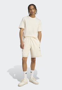 Kremo spalvos medvilninis marškinėliai, derantys šortai su elastine juosta. Baltos kojinės ir kreminės spalvos sportbačiai su akcentais. Paprastas logo detalė.