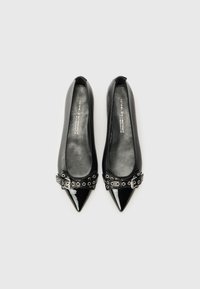 Ballerines en cuir noir avec un bout pointu, présentant un accent en vernis noir brillant et une ceinture décorative avec des œillets en argent et un détail de boucle.