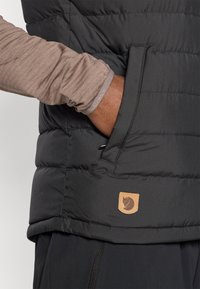 Zwarte gewatteerde bodywarmer met een voorvak en een tan kleurig logo-label. Het materiaal heeft een gestructureerde uitstraling met een gladde afwerking en zichtbare stiksel details.