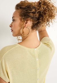 Haut à manches courtes jaune clair avec une finition texturée, présentant un col rond et un détail de bouton dans le dos. De grandes boucles d'oreilles en anneau sont portées.