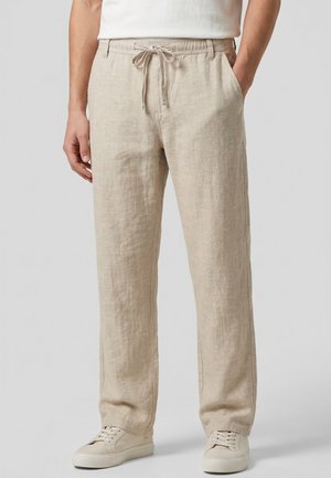 Beige Leinenhose mit Kordelzugbund, getragen mit weißen Sneakers und einem weißen T-Shirt, Hände teilweise in den Taschen.