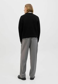 Osoba se středně dlouhými blond vlasy, oblečená v černém svetru, šedých širokých kalhotách a černých botách, stojí zády k bílé zdi.