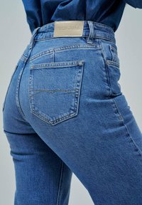 Vaqueros de denim azul con un diseño ajustado. Las características incluyen una etiqueta de cuero en la cintura, bolsillos traseros con detalles de costura y una textura suave.