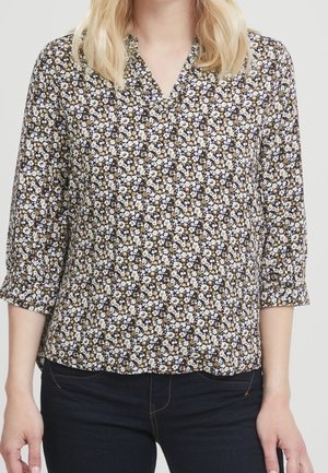 Femme portant un chemisier noir à fleurs avec de petites fleurs blanches et beiges, manches trois-quarts, et un jean bleu foncé.