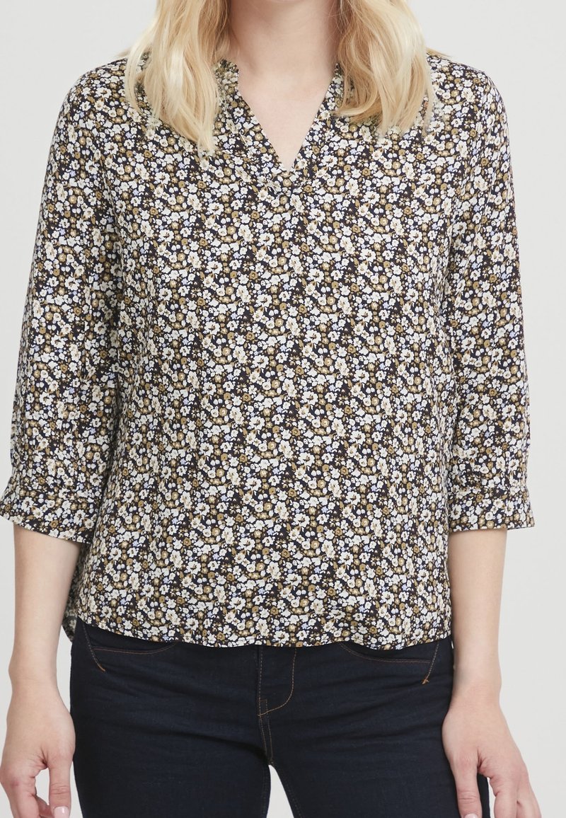 Femme portant un chemisier noir à fleurs avec de petites fleurs blanches et beiges, manches trois-quarts, et un jean bleu foncé.