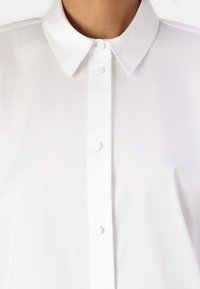 Camicia bianca con colletto classico e chiusura frontale a bottoni. Tessuto liscio senza fantasie; progettata per un aspetto pulito e minimalista.