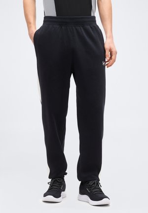 Άνδρας που φοράει μαύρο φόρμα με πλαϊνές τσέπες, ελαστική μέση και μαύρα αθλητικά παπούτσια με λευκές σόλες, σε απλό φόντο.