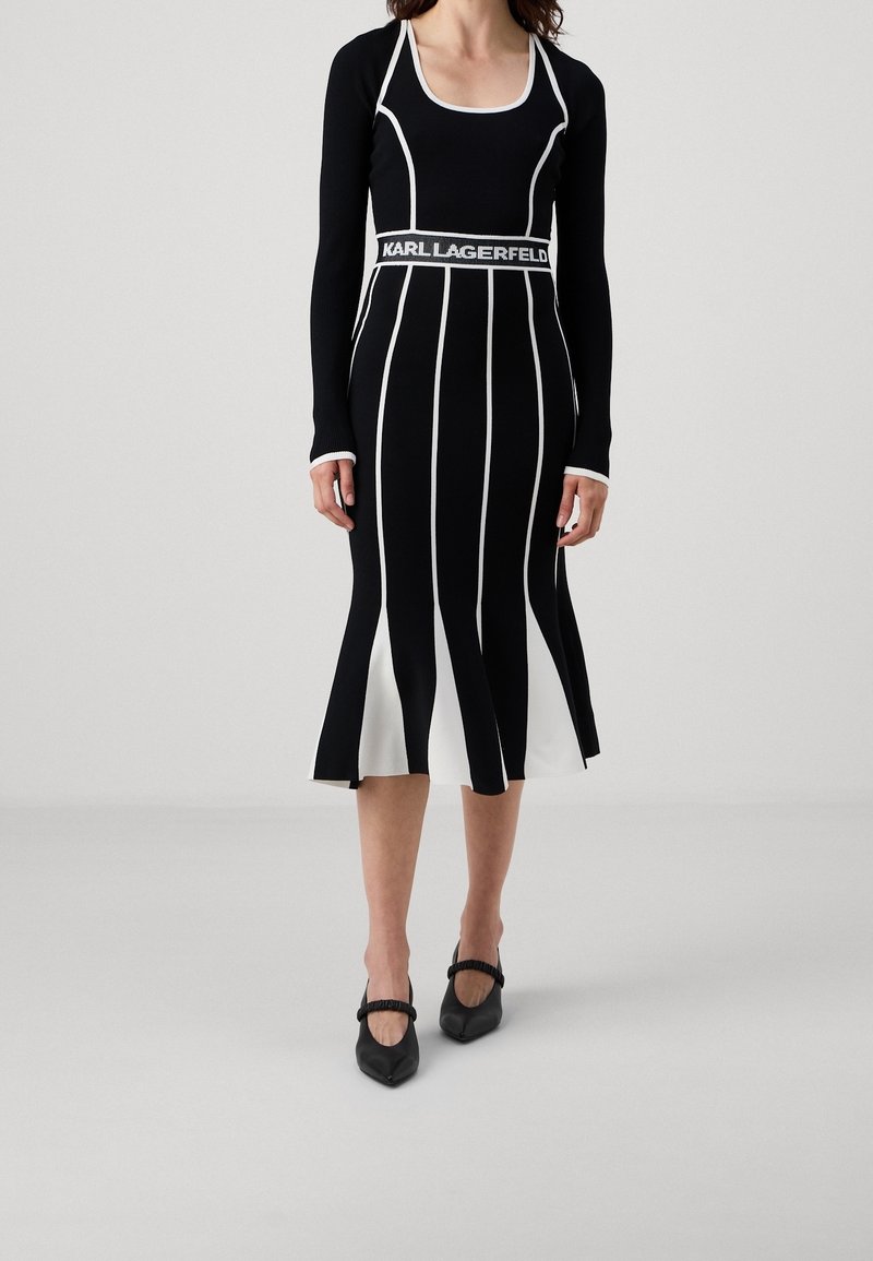 Femme portant une robe noire ajustée à manches longues avec des rayures verticales blanches et le texte "Karl Lagerfeld" à la taille, associée à des chaussures noires pointues.