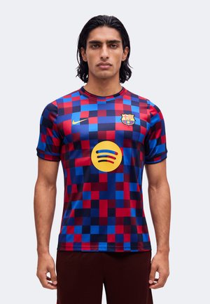 Homme portant un maillot de football à carreaux bleu et rouge du FC Barcelone avec les logos Nike et Spotify, debout devant un fond clair uni.