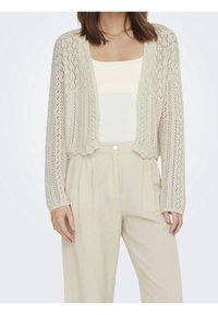 Cárdigan corto de punto en crema claro, con un patrón de encaje, abertura frontal y mangas largas. Lleva puesto sobre una camisa blanca y pantalones beige.