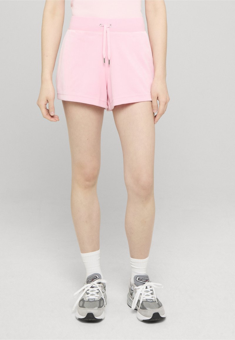 Shorts roses en matériau doux, avec une taille à cordon de serrage et une coupe décontractée, assortis à des baskets grises et des chaussettes montantes blanches.