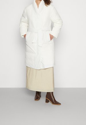 Woman wearing a white belted puffer coat over a beige midi skirt and dark brown heeled ankle boots, standing against a white background.
