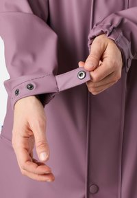 Paarse waterdichte jas manchette met drukknoopsluiting. Materiaal is glad en flexibel. Zichtbare hand die de mouw aanpast.