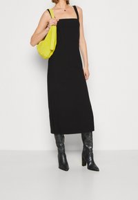 Robe midi noire sans manches avec un décolleté carré ; associée à un sac en daim jaune et des bottes noires texturées jusqu'aux genoux.