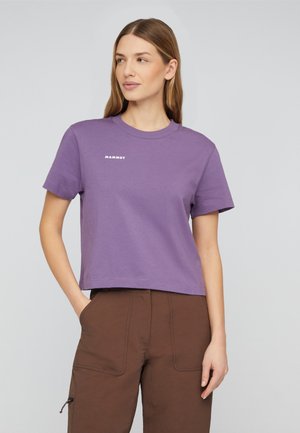 Kvinde iført en lilla Mammut t-shirt og brune bukser, stående med den ene hånd i lommen, kigger let til siden.