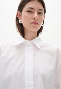 Bílá košile s límečkem vyrobená z hladké látky, s předním zapínáním na knoflíky a oversized střihem. Stříbrné kulaté náušnice dotvářejí vzhled.