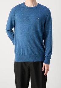 Niebieski sweter dziany z nakrapianą teksturą. Rękawy i dół wykończone ściągaczem, okrągły dekolt. Małe logo na lewej piersi. Czarne spodnie.