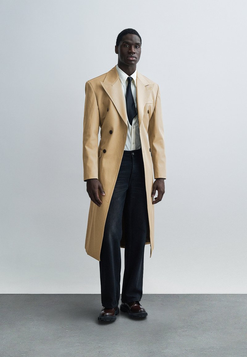 Trench-coat beige croisé avec de larges revers, porté sur une chemise blanche et une cravate noire. Un jean foncé et des chaussures marron complètent le look.