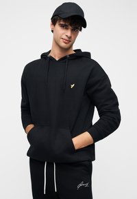 Μαύρο φούτερ με κουκούλα και χρυσό λογότυπο πουλιού, με τσέπη μπροστά και κορδόνια. Συνδυάζεται με αντίστοιχο μαύρο παντελόνι φόρμας.