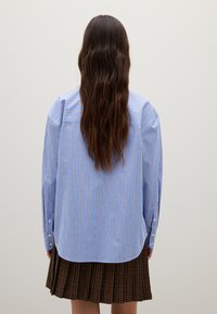 Blue and white striped shirt with long sleeves and button cuffs, featuring a curved hem and a relaxed fit, paired with a pleated skirt.