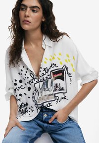 Weißes Hemd mit Knopfleiste, verziert mit abstrakten Mustern in Schwarz und Gelb sowie einem Grafikdesign auf der Vorderseite. Kombiniert mit lockeren blauen Jeans.