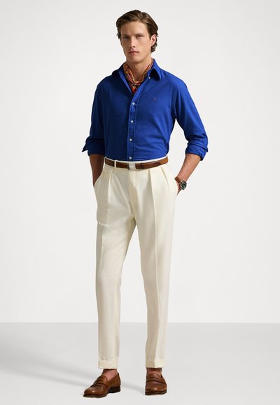 Blue button-up shirt with long sleeves, cream pleated trousers, brown leather belt, and brown loafers. Silk patterned scarf accents the neck.