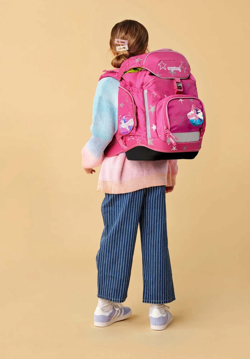 Kind trägt einen pastellfarbenen Pullover und gestreifte Hose, trägt einen großen rosa Rucksack mit Stern- und Pferdemotiven, blickt auf einem beigen Hintergrund weg.