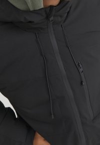 Černá bunda s kapucí, která má plný zip, nastavitelné šňůrky a texturovanou látku. Dezén švů a minimální kovové akcenty.