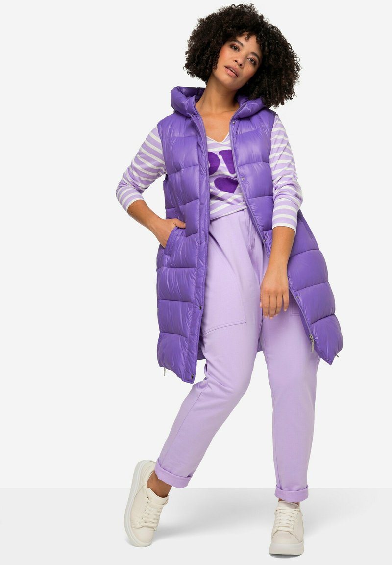 Gilet rembourré violet avec capuche, associé à un t-shirt à manches longues rayé violet et blanc et un pantalon violet clair. Baskets blanc cassé.