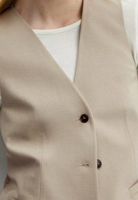 Beżowa bezrękawnik uszyty z teksturowanej tkaniny, z głębokim dekoltem w kształcie litery V, dwoma ciemnymi guzikami i minimalistycznymi akcentami wzorniczymi.